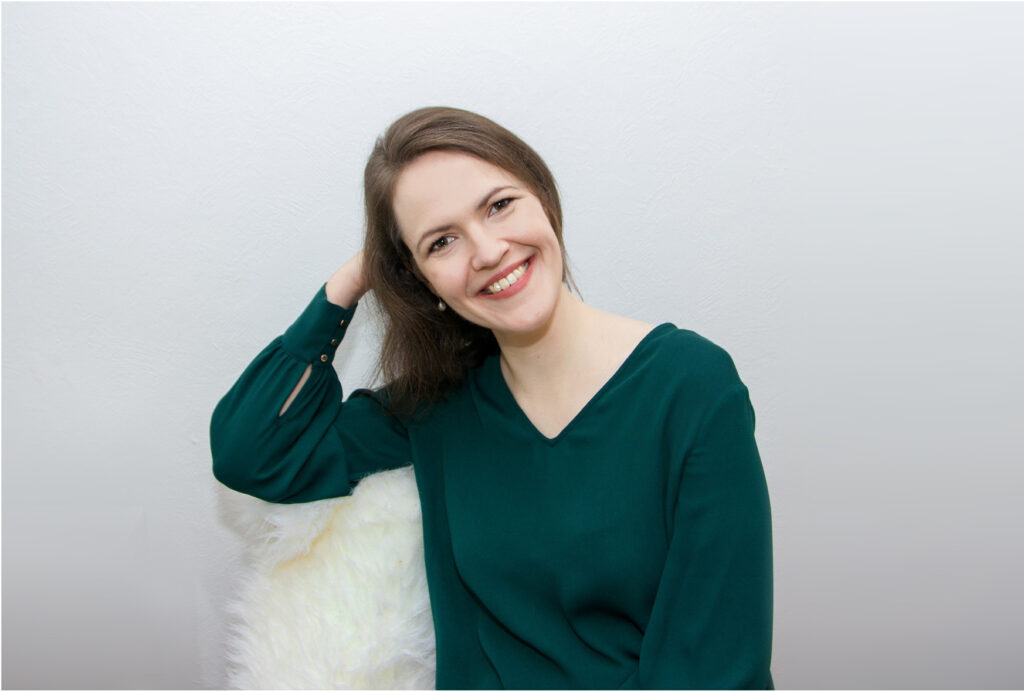 Profilbild von Anna Augustin, Frau mit braunen Haaren und dunkelgrüner Bluse, Kopf auf rechte Hand gestützt