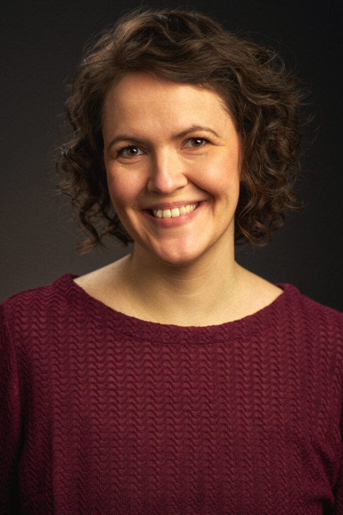 Über mich Profilbild von Anna Augustin, Frau mit braunen Haaren und dunkelroter Bluse, lachend