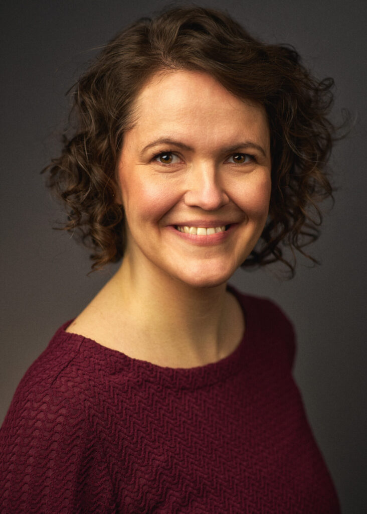 Über mich Profilbild von Anna Augustin, Frau mit braunen Haaren und dunkelroter Bluse, lachend