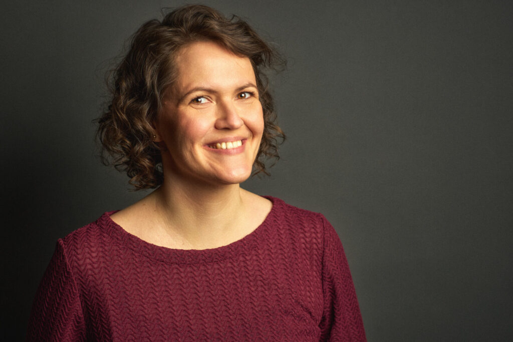Startseite Profilbild von Anna Augustin, Frau mit braunen Haaren und dunkelroter Bluse, lachend