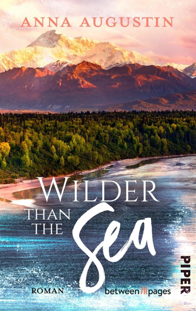 Cover des Romans Wilder than the Sea von Anna Augustin, Ozeanküste vor einem dichten Wald, im Hintergrund ein Bergmassiv vom Sonnenuntergang beleuchtet, rosafarbener Himmel